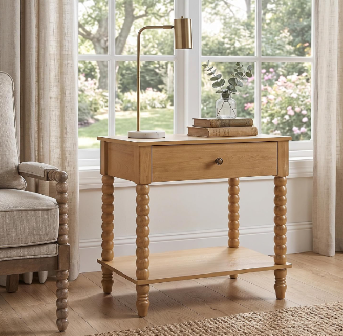 A brown wood side table with a single drawer, turned spindle legs, and a lower shelf, styled in a bright room next to a window with beige curtains.