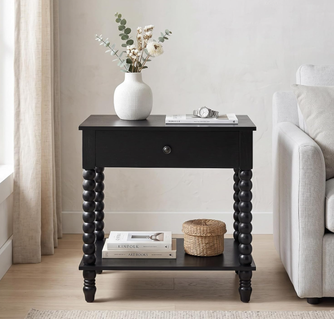 A black wooden side table with a single drawer and open lower shelf, styled with a white vase of flowers, books, and a small basket, set in a bright living room next to a light gray sofa.